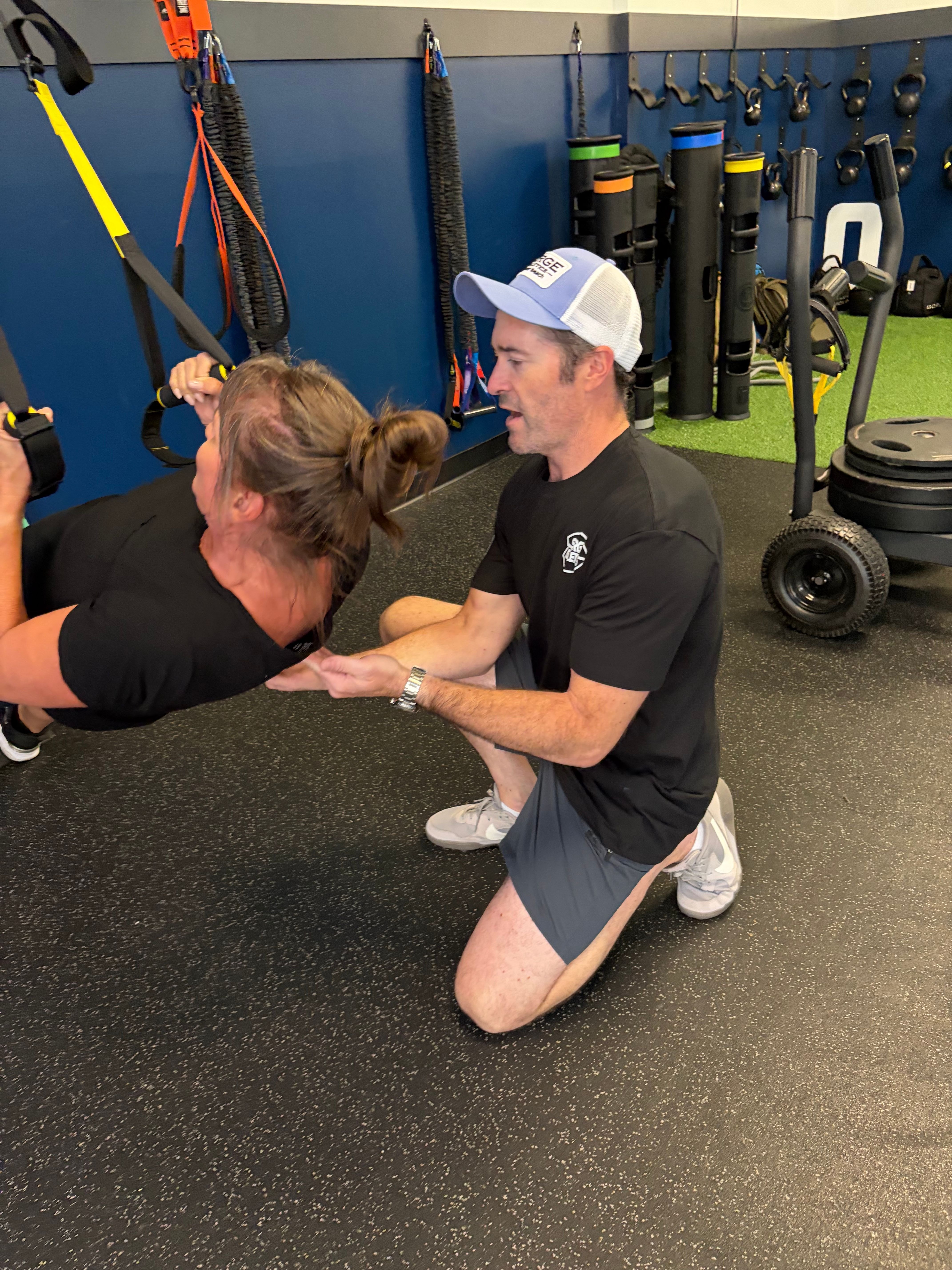 Trainer in a blue cap kneeling to guide a woman through a suspension trainer exercise at Sarge Athletics