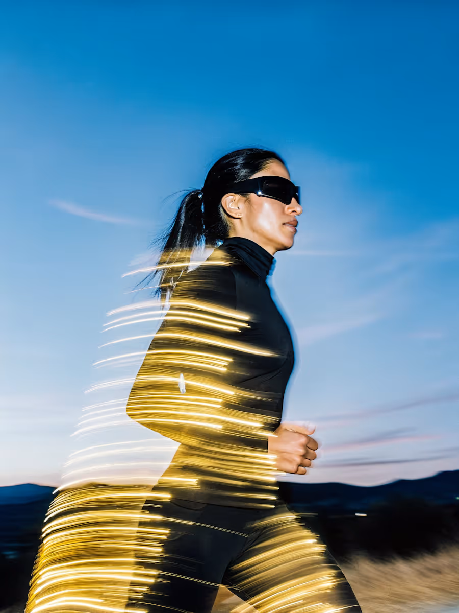 Woman in black athletic wear and sunglasses running outdoors at dusk with yellow light trails around her body.