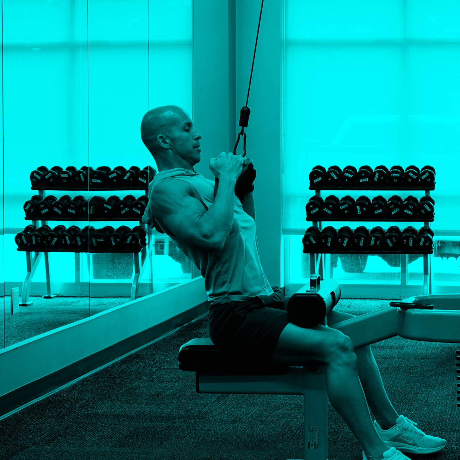 Man doing seated cable row exercise in a gym with a rack of dumbbells and mirrored wall in the background.