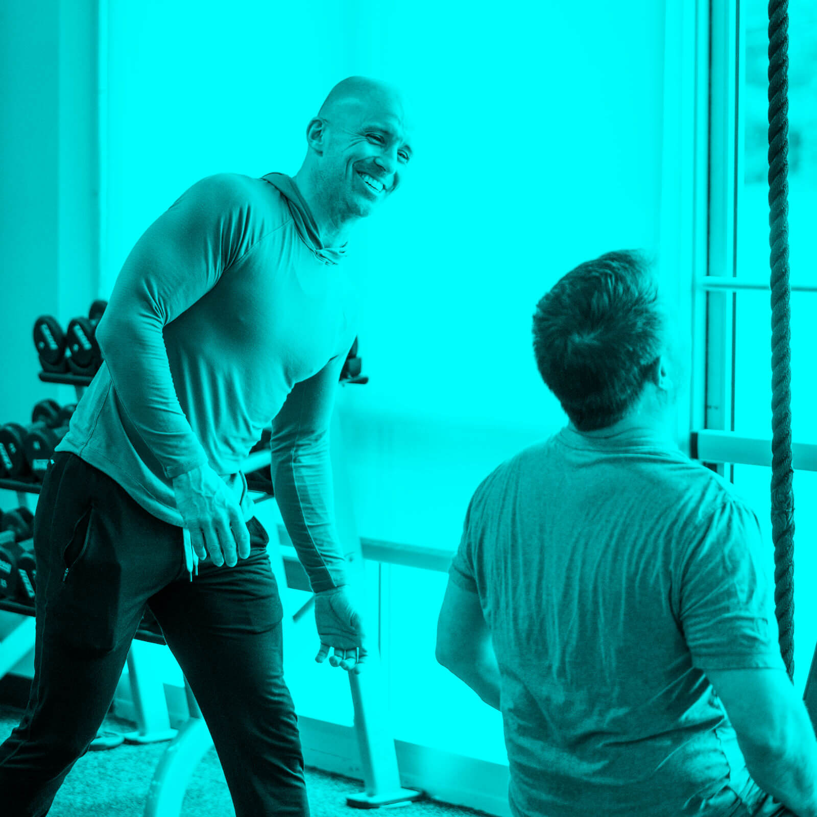 Fitness coach smiling and assisting a man working out on gym equipment near a window.