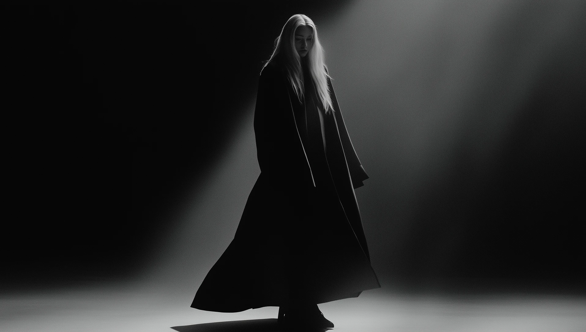 Model with long light hair wearing a structured black coat, standing in a minimalist dark space illuminated by a soft directional spotlight.