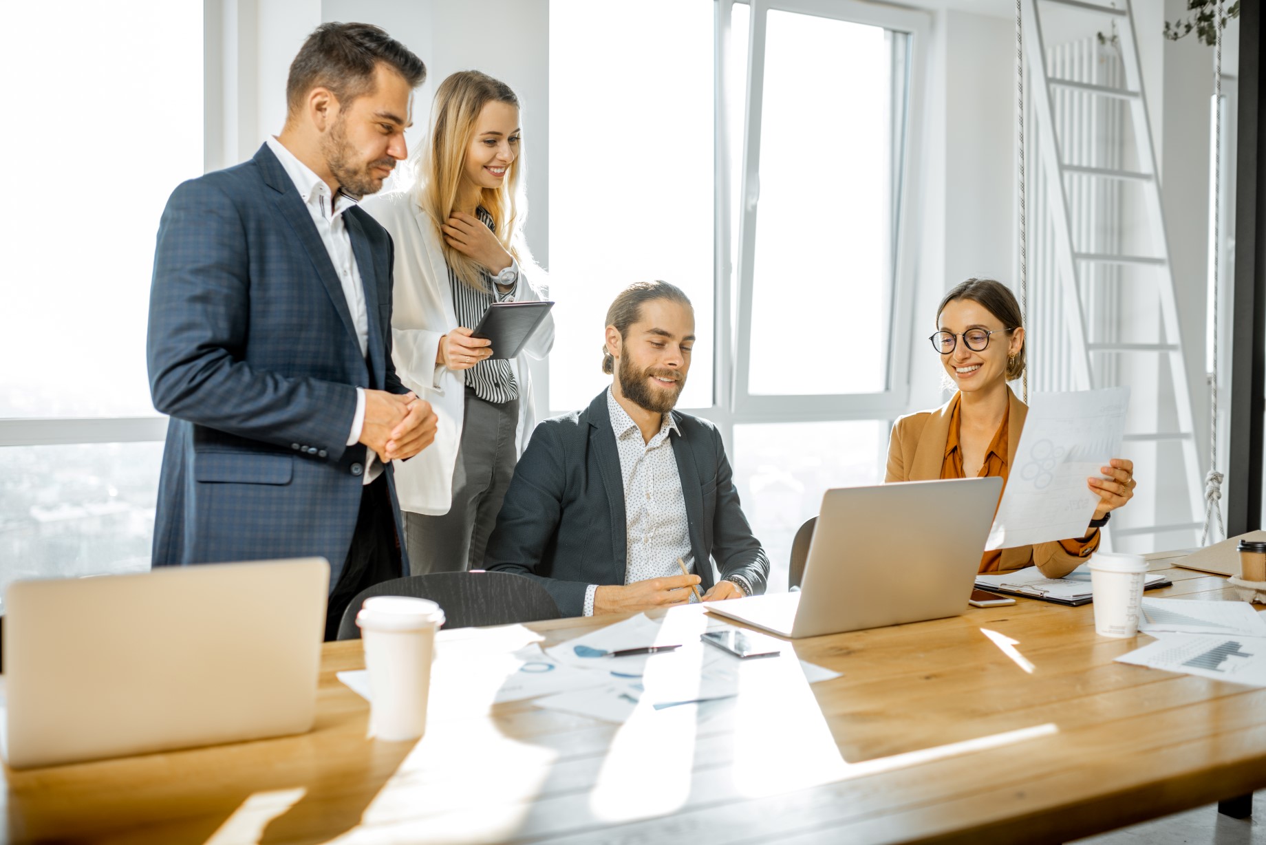 Czterech pracowników w jasnym biurze, dwóch stojących i dwóch siedzących przy biurku, którzy omawiają projekt.