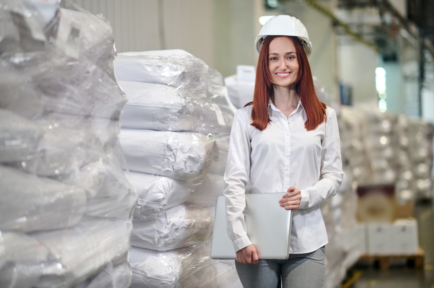 Kobieta w białym kasku na tle wnętrza zakładu produkcyjnego.