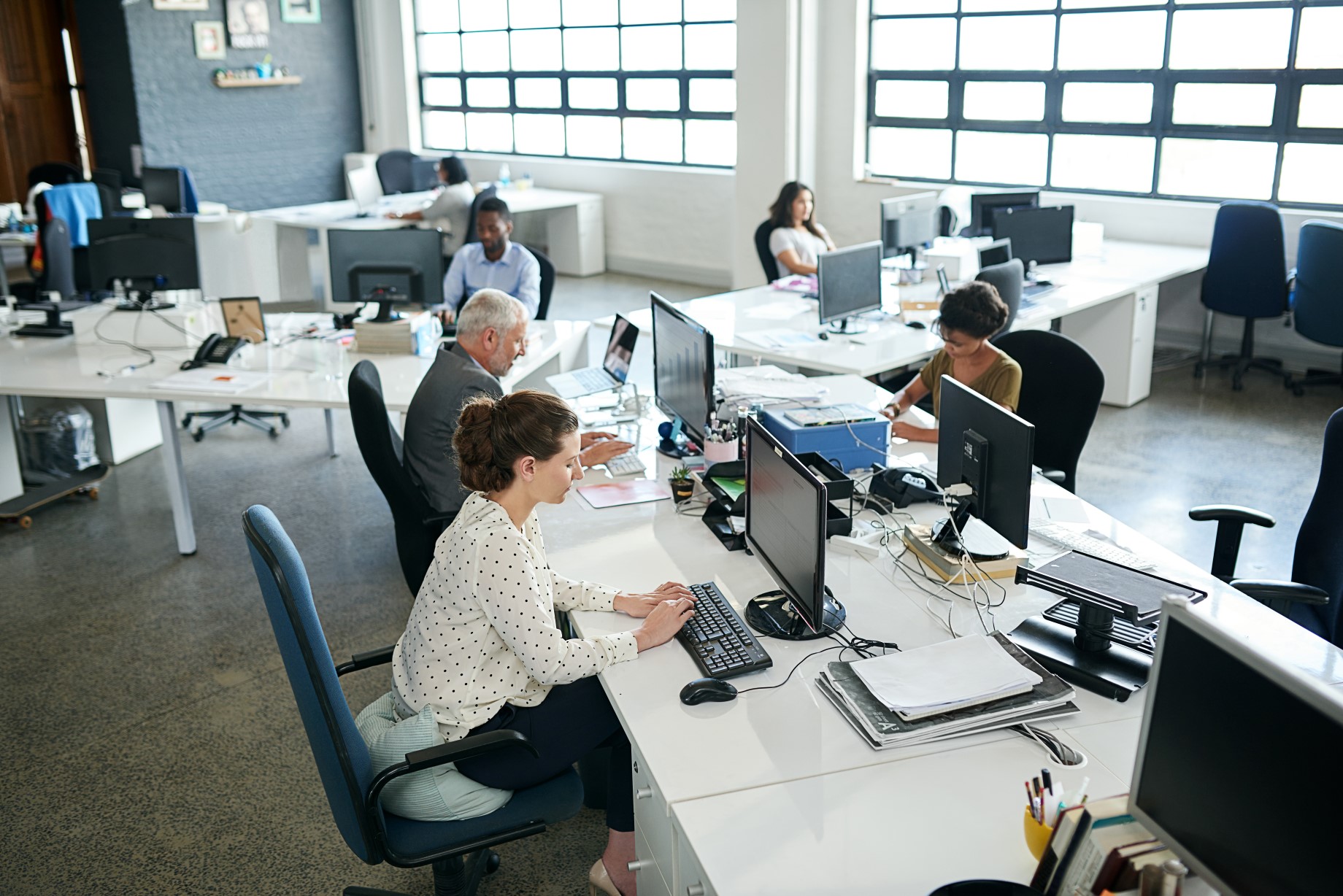 Przestronne biuro z dużymi oknami. Grupa pracowników przy biurkach, pracujących na komputerach.