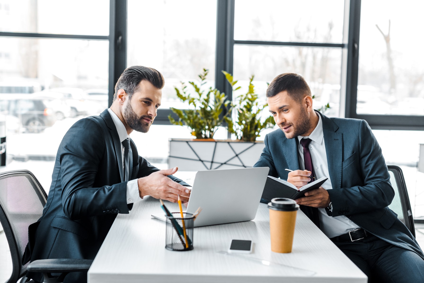 Dwóch mężczyzn w garniturach siedzących w biurze przy biurku naprzeciwko siebie 