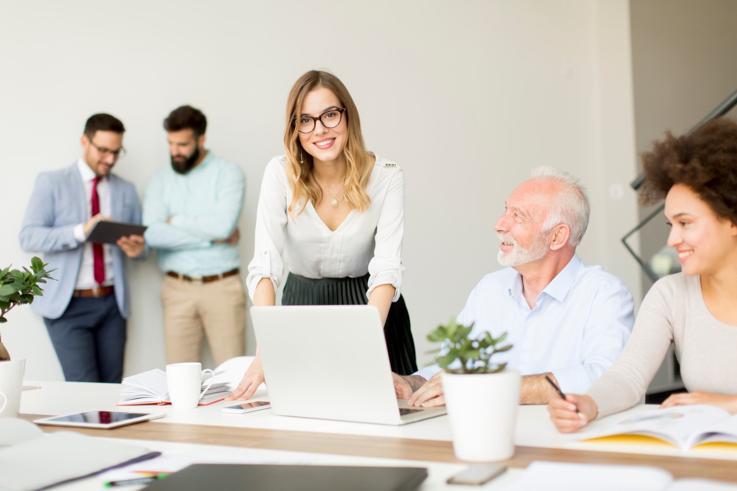 Pracownicy w dobrze oświetlonym biurze. Dwie osoby siedzą przy biurku z otwartym laptopem, elegancka kobieta stoi pochylona nad biurkiem i uśmiecha się.
