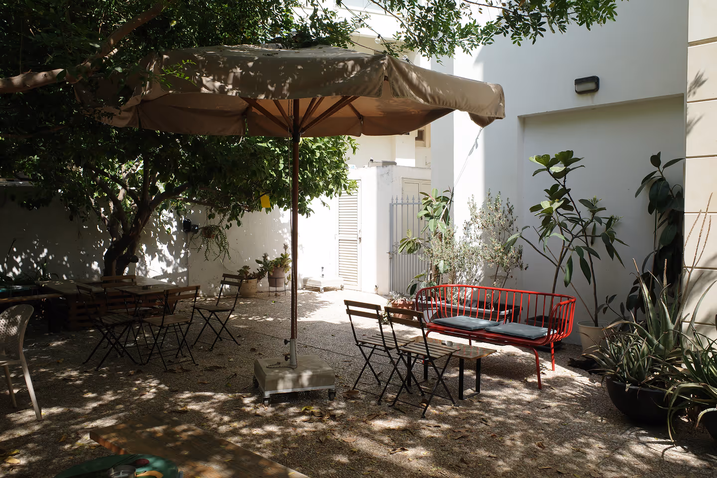 An outdoor courtyard shaded by trees and a large umbrella. There are simple wooden tables and chairs, a red bench, and lots of plants.