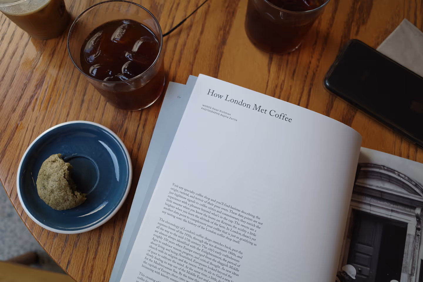 A wooden table set with two glasses of cold brew, a blue plate holding a partially eaten matcha cookie, and an open magazine with an article titled 'How London Met Coffee.