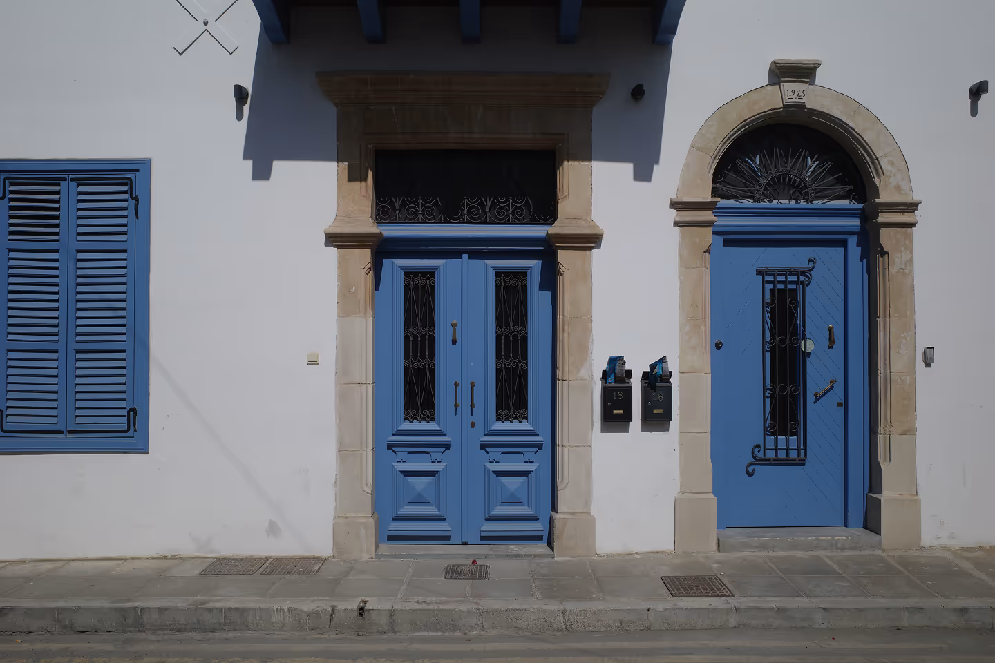 A street view of a white building with two blue doors and matching shutters. The building has an architectural blend of modern and classic styles, with stone framing around the doors, and a plaque above one door marked 1925.