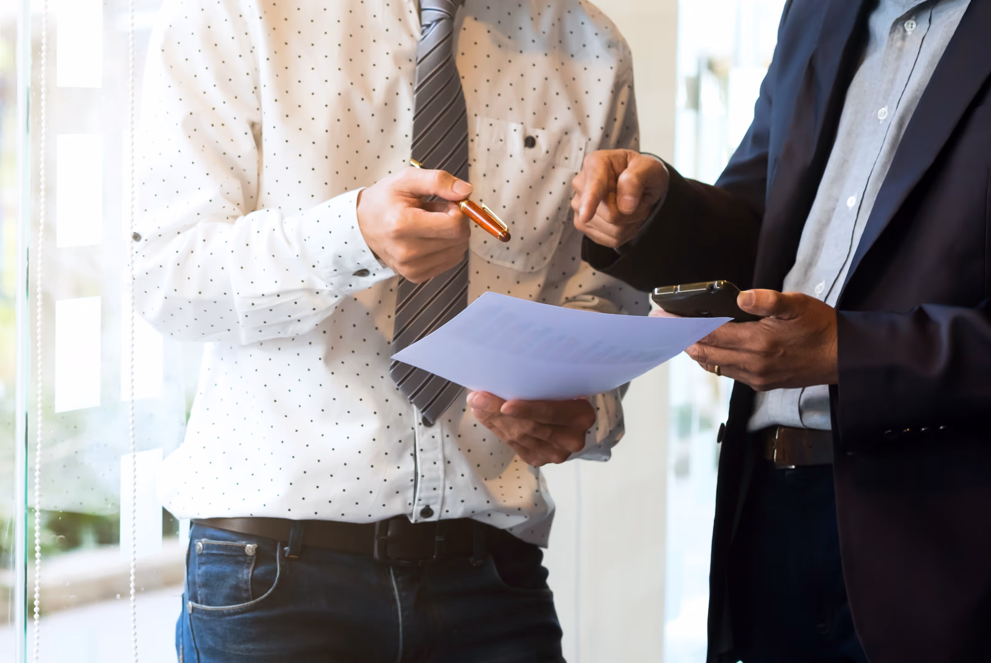 Dos hombres de negocios revisando un documento, uno sostiene una hoja y bolígrafo y el otro un teléfono móvil.