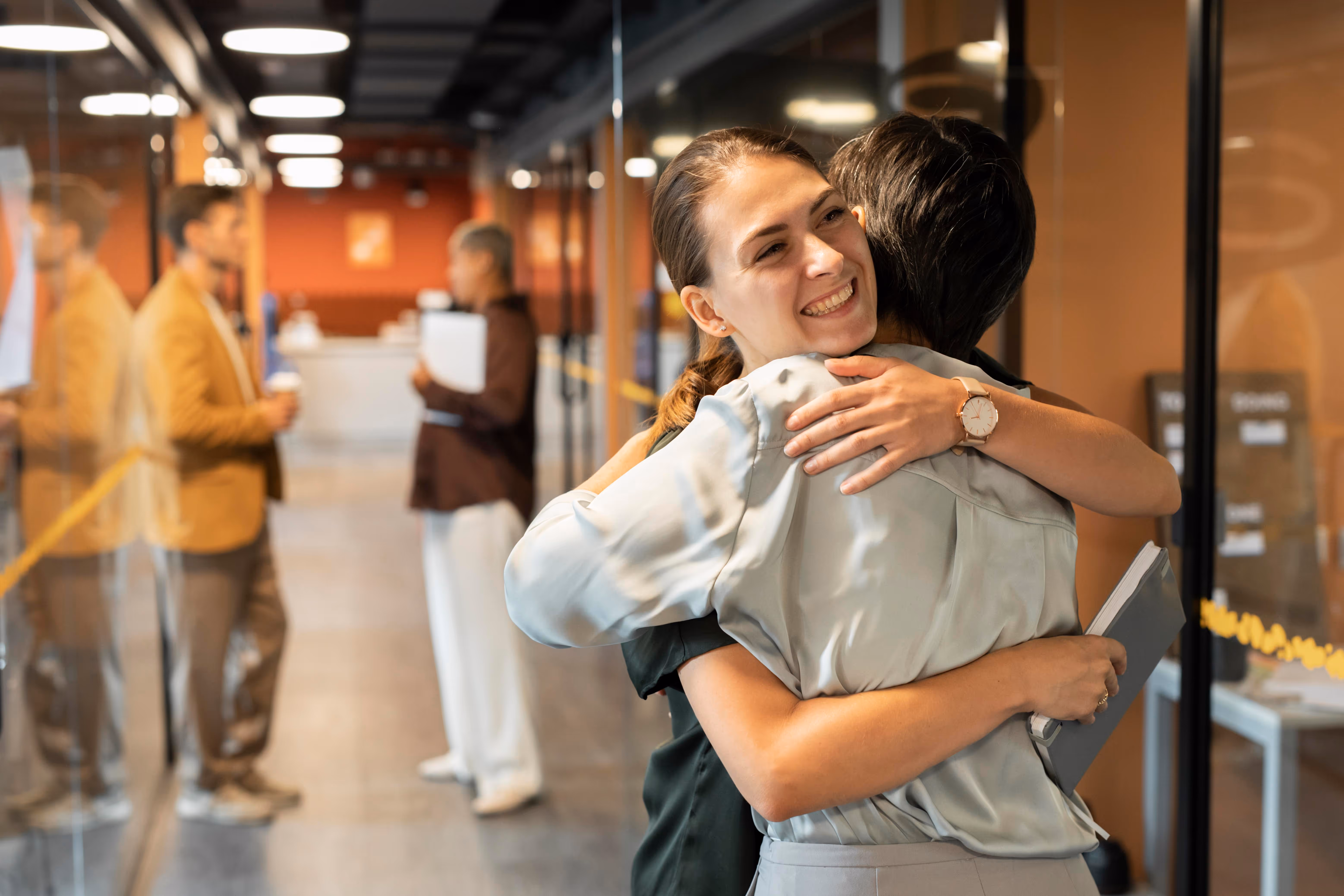 Dos compañeras de trabajo abrazándose con sonrisas en un pasillo de oficina moderno.