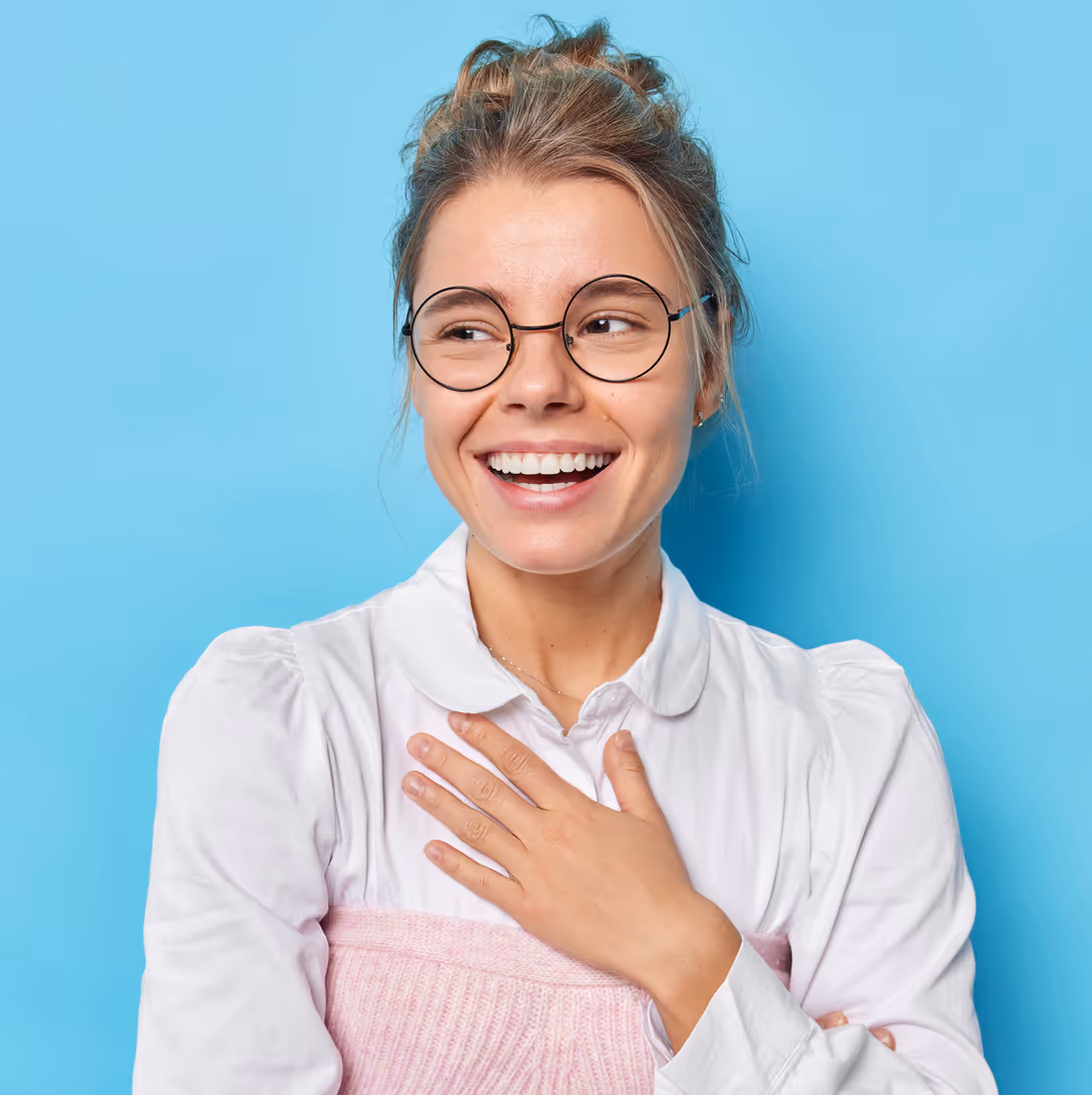Mujer sonriente con gafas redondas y mano sobre el pecho frente a fondo azul.
