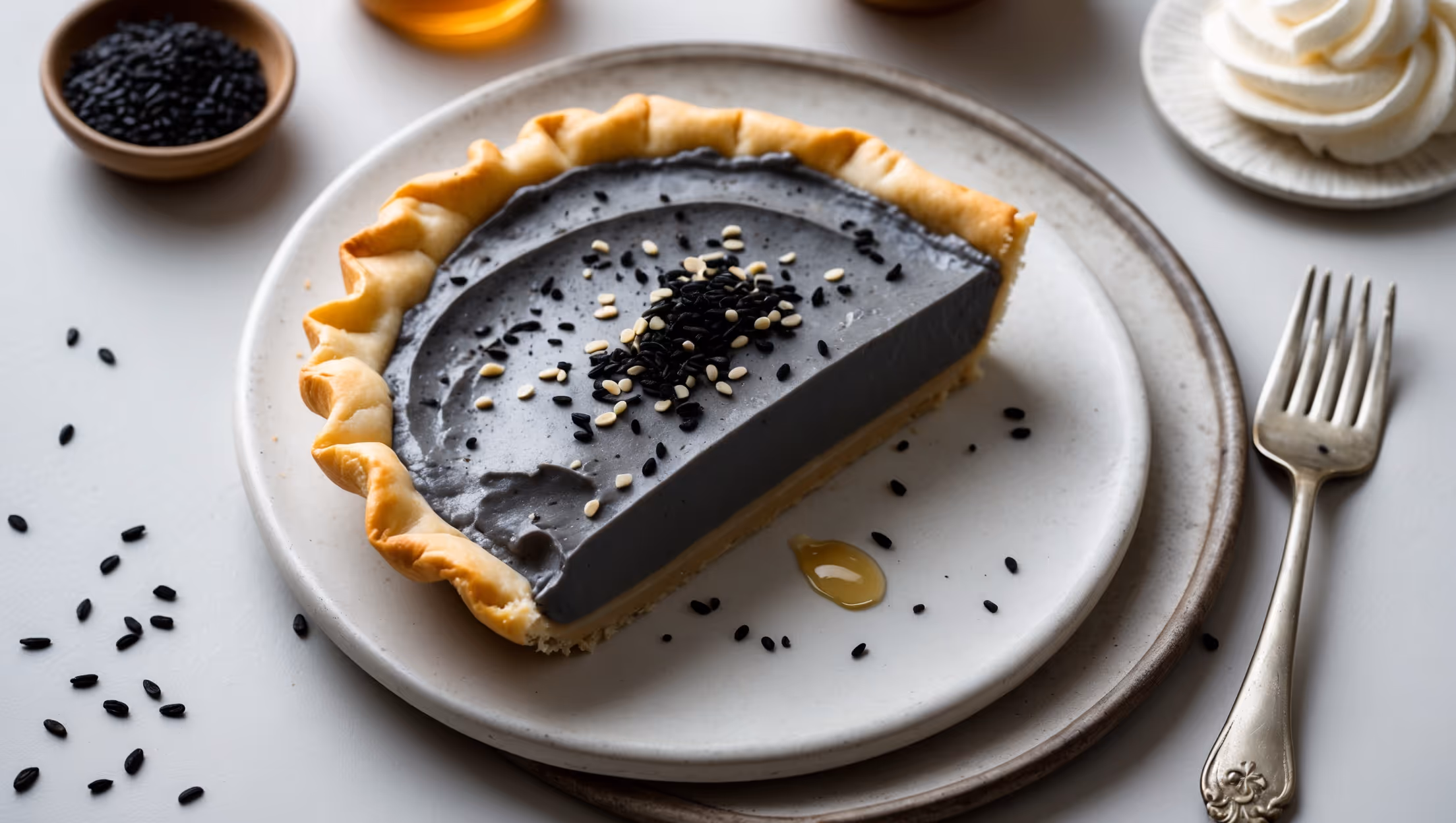 An overhead shot of a slice of Black Sesame Cream Pie revealing its striking dark gray filling against a golden brown pastry crust. Soft natural light accentuates the velvety surface texture of the cream filling, which is garnished with a sprinkle of black sesame seeds and delicate white sesame seeds creating visual contrast. The pie sits on a minimalist white ceramic plate with a vintage silver dessert fork beside it. In the background, a small bowl of whole black sesame seeds and a drizzle of honey suggest the key flavors. The cross-section reveals the perfect layers: flaky crust, smooth sesame filling, and cloud-like whipped cream topping that's been artfully piped.