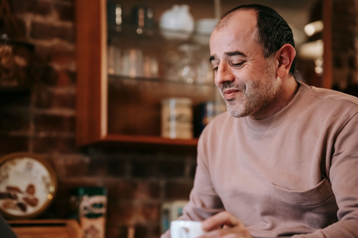 A man sits at a kitchen table with a cup and smiles as he looks down