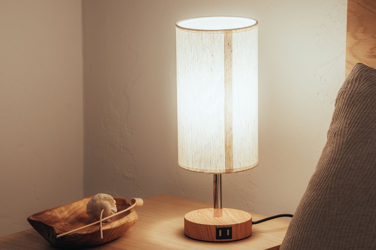 A lit table lamp on a wooden nightstand beside a bed