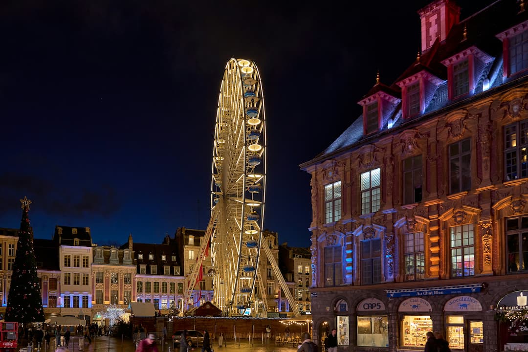 Grande Roue illuminée et marché de Noël sur une place animée de Lille la nuit, avec des bâtiments historiques éclairés.