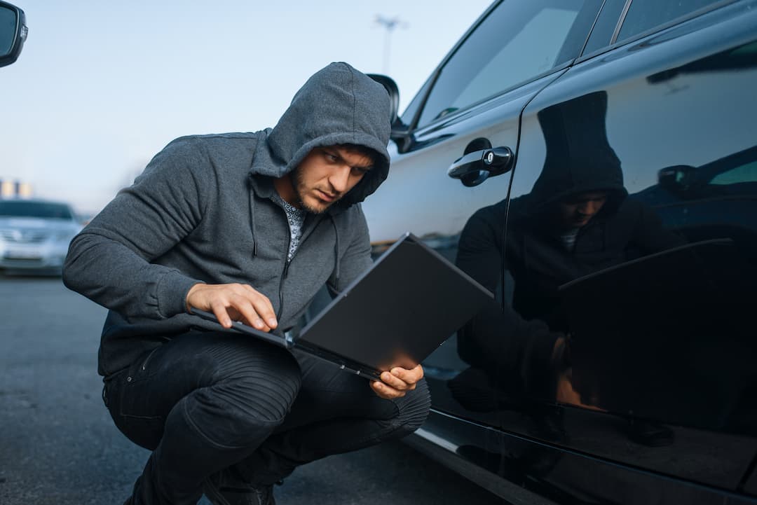 Un pirate informatique en sweat à capuche utilise un ordinateur portable pour contourner le système d'une voiture, illustrant les risques d'arnaque pour vendeur automobile via la technologie.