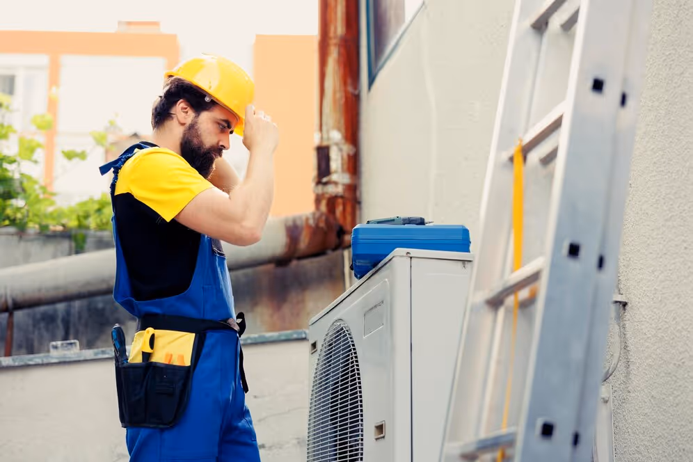 Worker in yellow hard hat and blue overalls adjusting helmet beside air conditioning unit and ladder