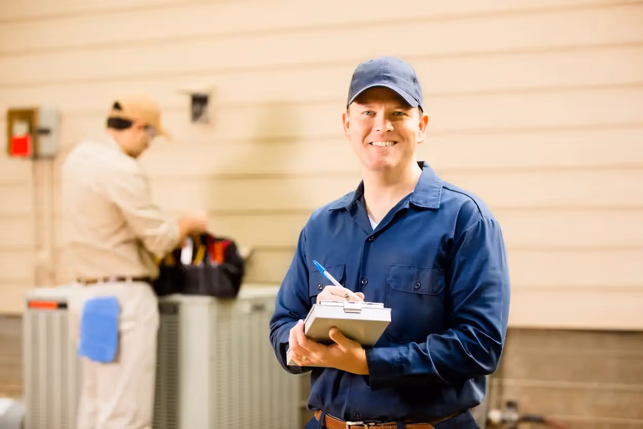 Smiling technician in blue uniform taking notes near HVAC equipment