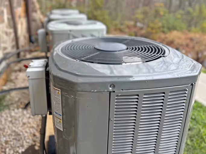 Row of gray outdoor air conditioning units with top fans placed on gravel surface beside building