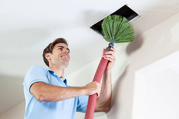 Person cleaning ceiling vent with long-handled green brush
