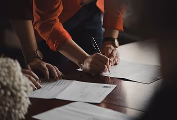 Person signing document with pen on wooden table, wearing red sleeve