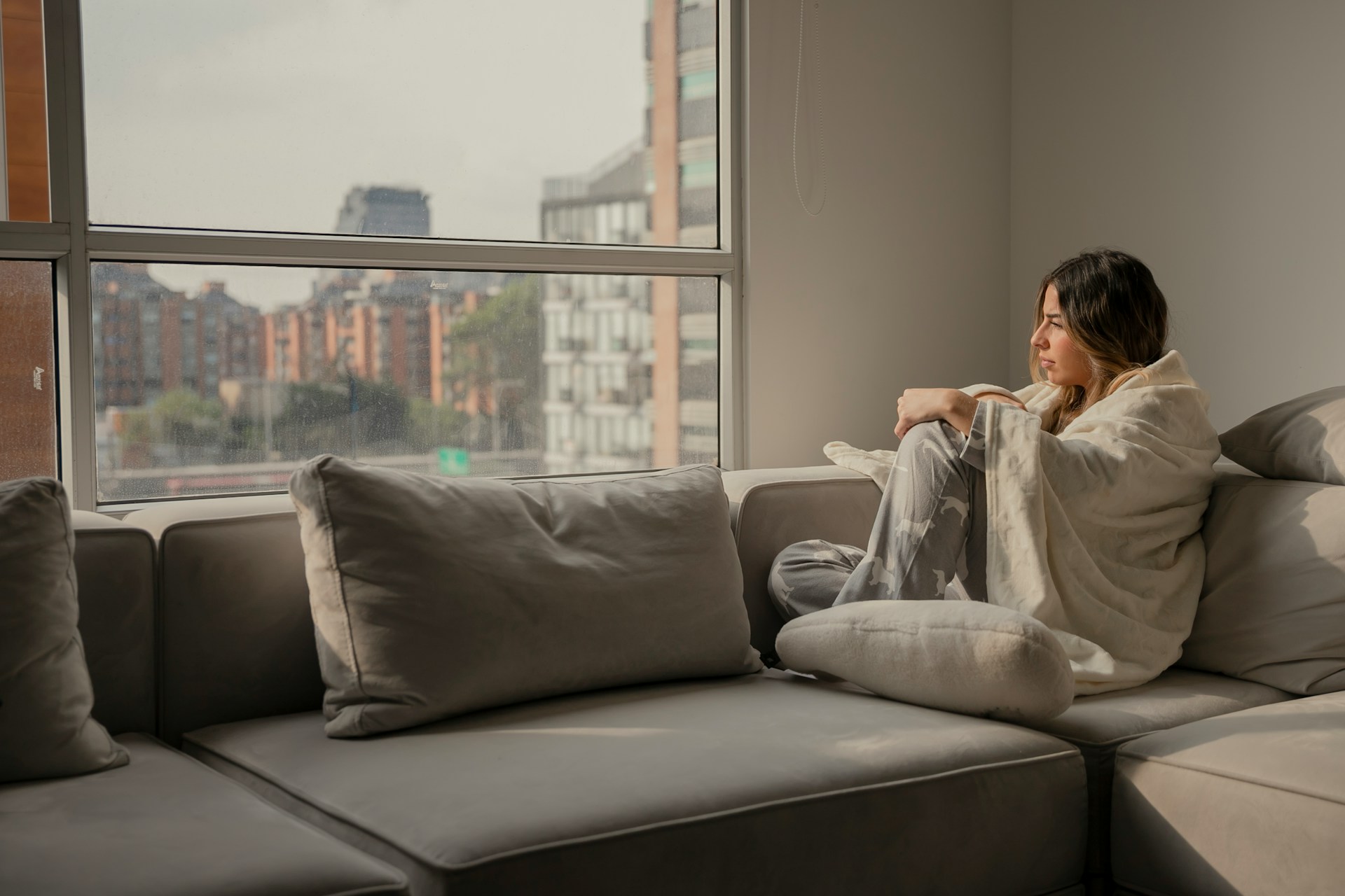 Person wrapped in blanket sitting on couch, looking out urban window