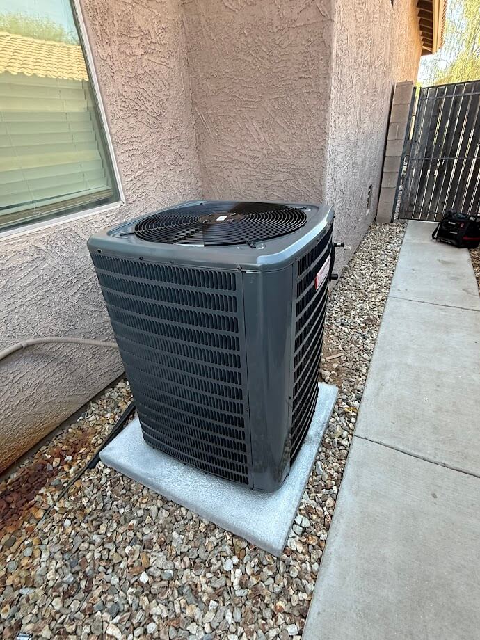 A large gray Goodman HVAC air conditioning unit sits on a concrete pad next to a house with stucco siding and a gravel landscape.