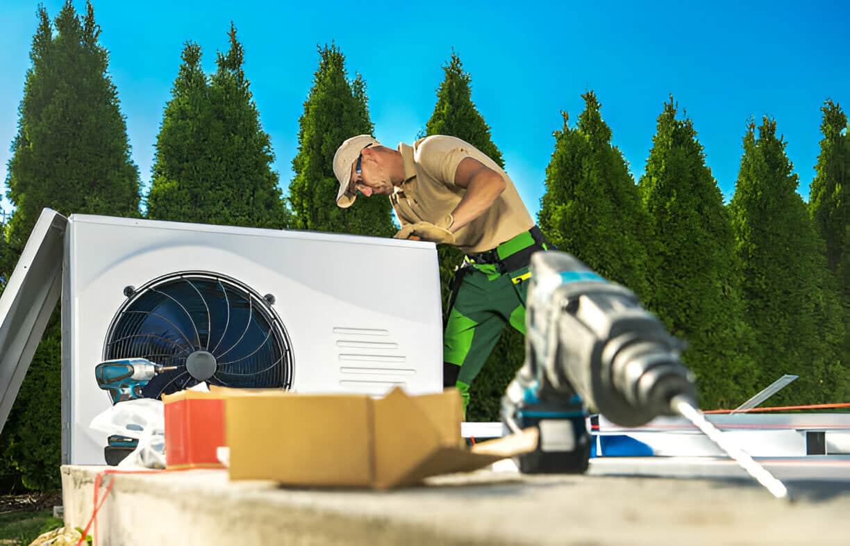 A service technician is installing or servicing an outdoor air conditioning unit, with a row of tall evergreen trees in the background.