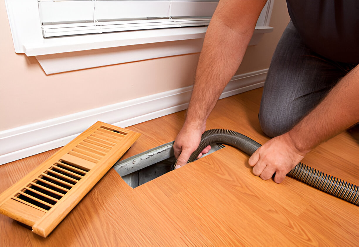 Person cleaning air vent with vacuum hose on wooden floor