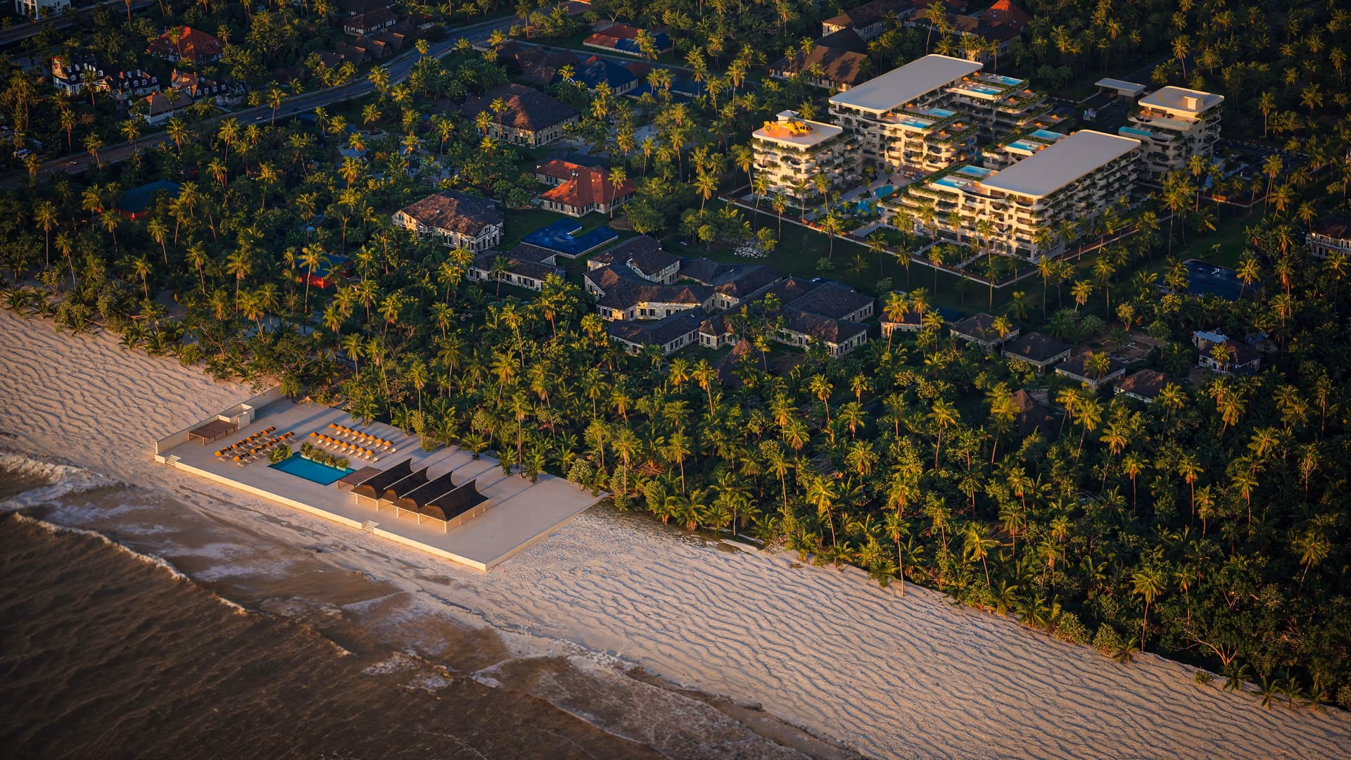 Aerial view of a beach resort with pools, sun loungers, palm trees, and modern buildings near the shoreline.
