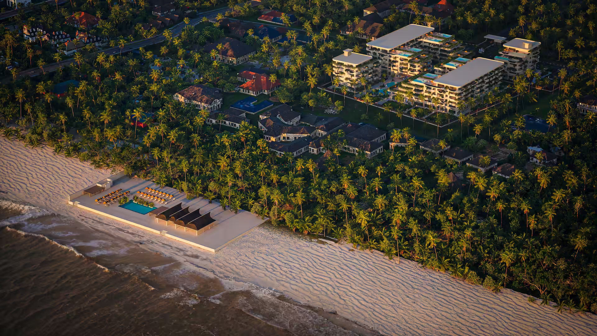 Aerial view of a beachfront resort area with sandy beach, an outdoor pool with lounge chairs, surrounding palm trees, various houses, and two modern multi-story buildings.