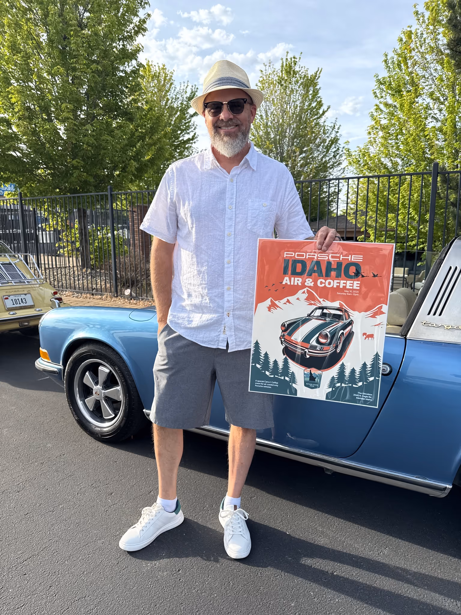 Air-cooled Porsche visitor holding Idaho Air & Coffee event poster next to a classic Porsche at Cars & Coffee in Idaho. 