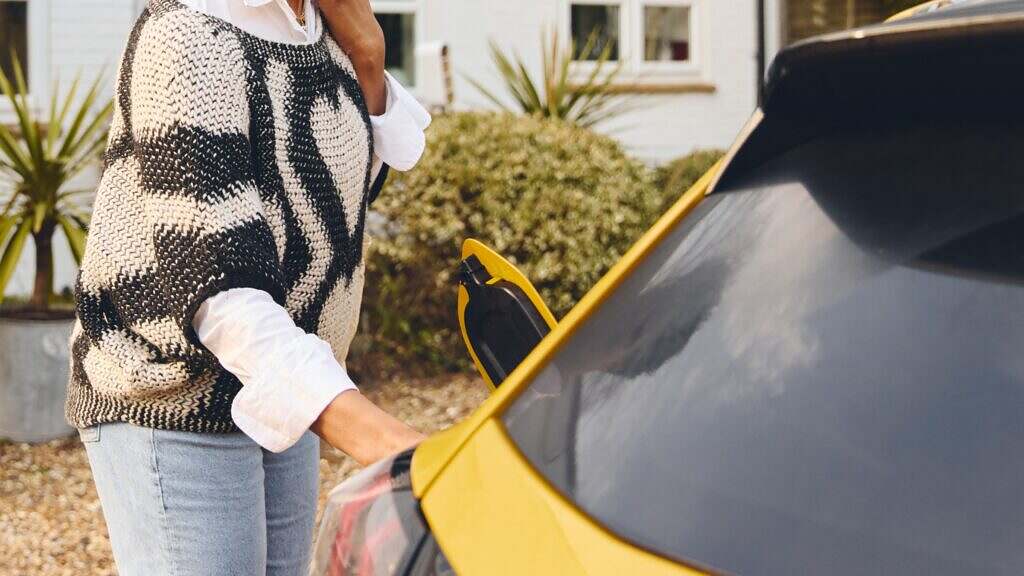 A person in a knitted sweater is standing by a yellow electric car. She has opened the charging hatch.