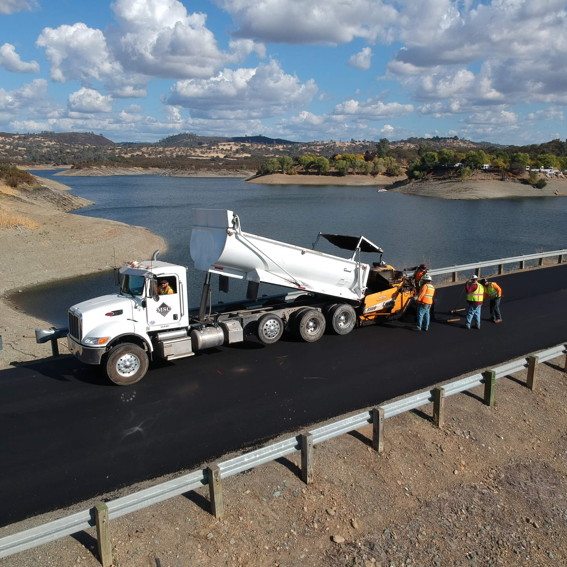 Workers in safety vests paving a road beside a lake, with a white dump truck and asphalt paving machine.