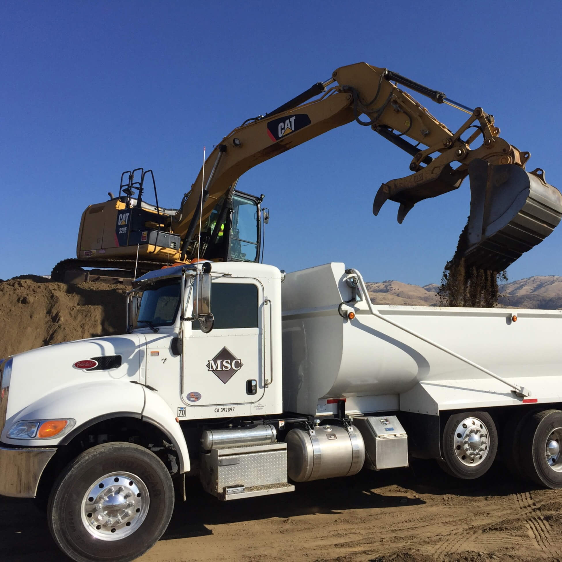 Yellow excavator loading dirt into the bed of a white MSC dump truck on a construction site with clear blue sky.
