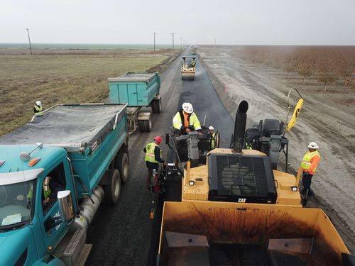 Construction workers in safety vests and helmets paving a road with heavy machinery and dump trucks in a rural area.