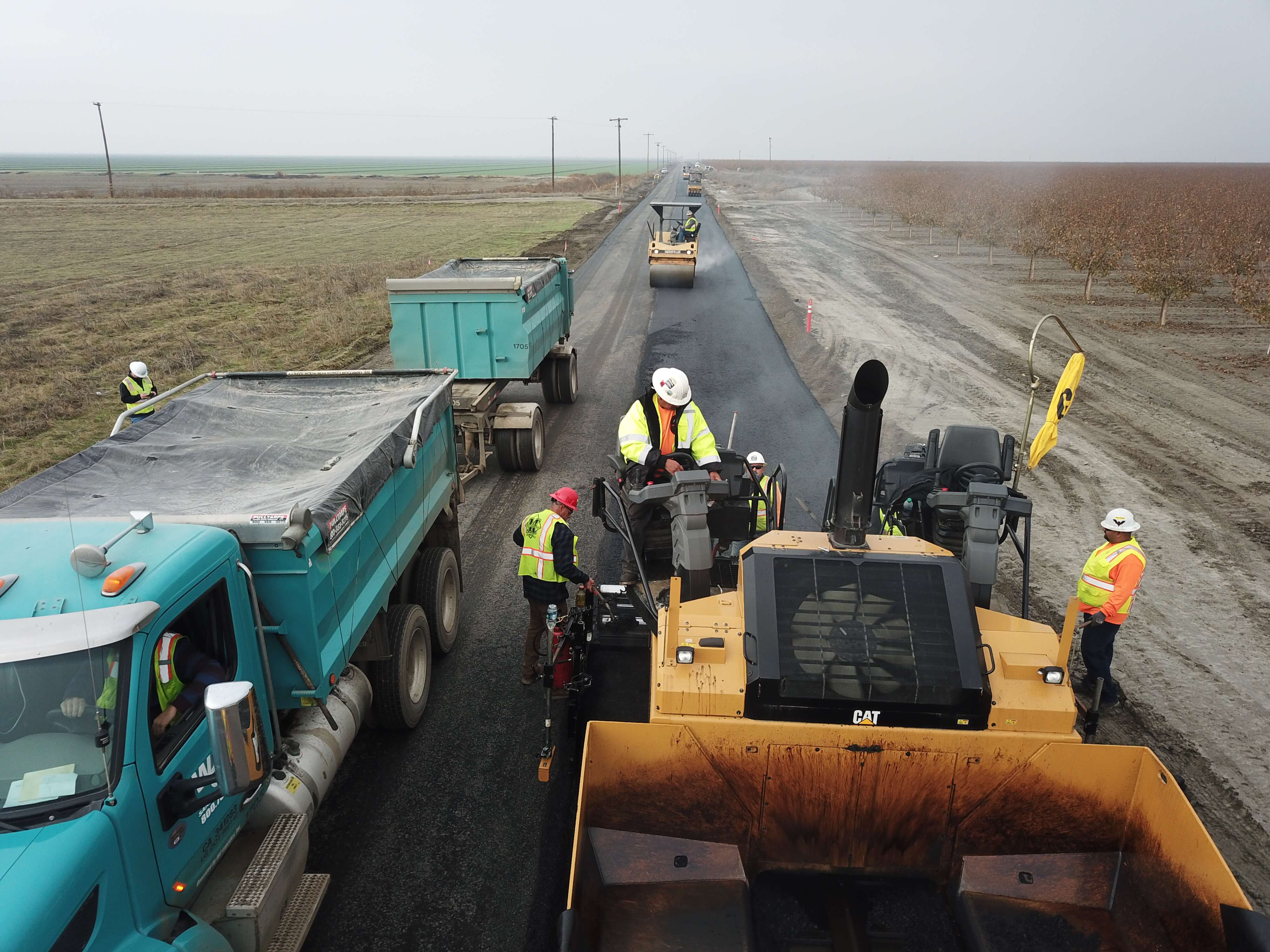 Construction workers in safety vests and helmets paving a road with heavy machinery and dump trucks in a rural area.