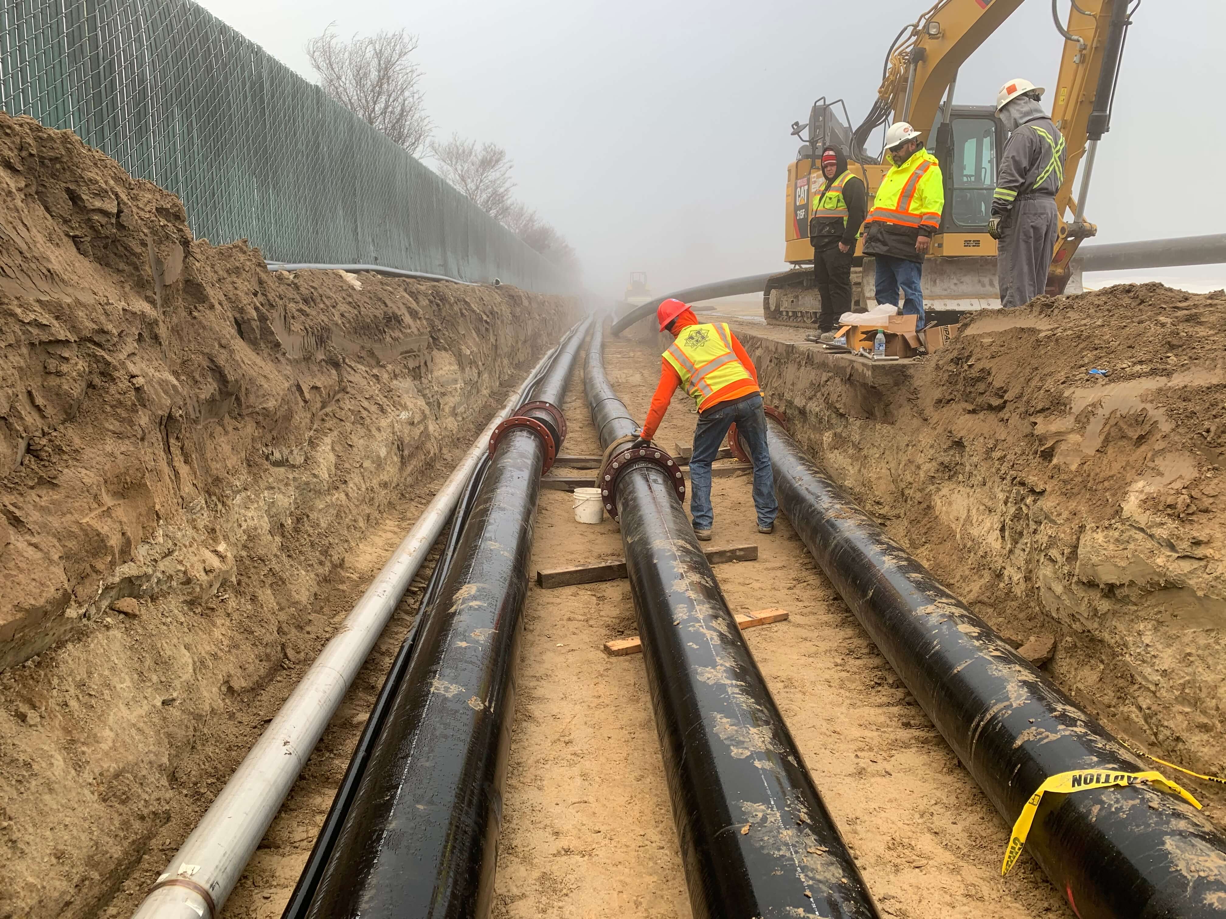 Construction workers in safety gear inspecting large black and metal pipes laid in a deep trench next to an excavator.
