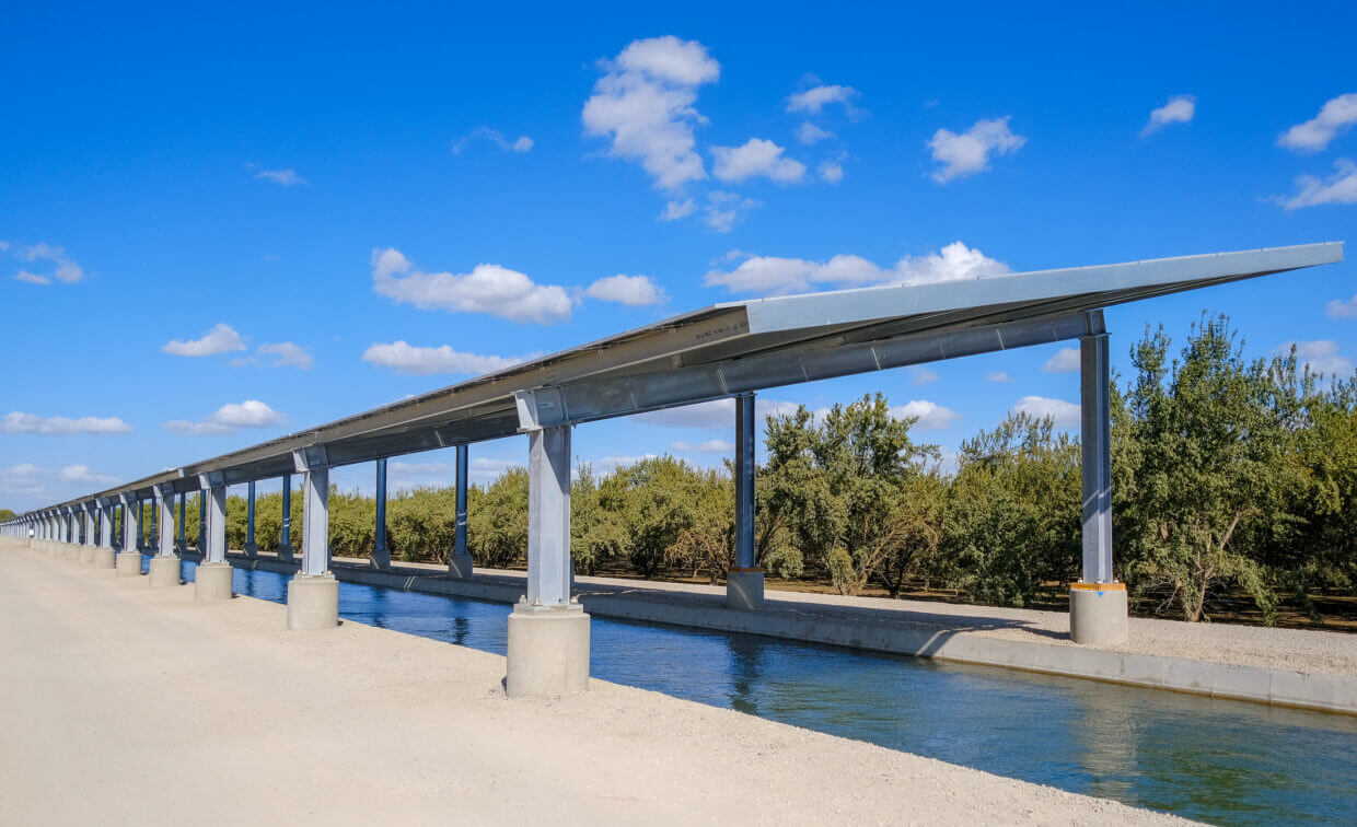 Long elevated aqueduct channel with metal supports over a concrete water canal next to a dirt path under a blue sky with scattered clouds.