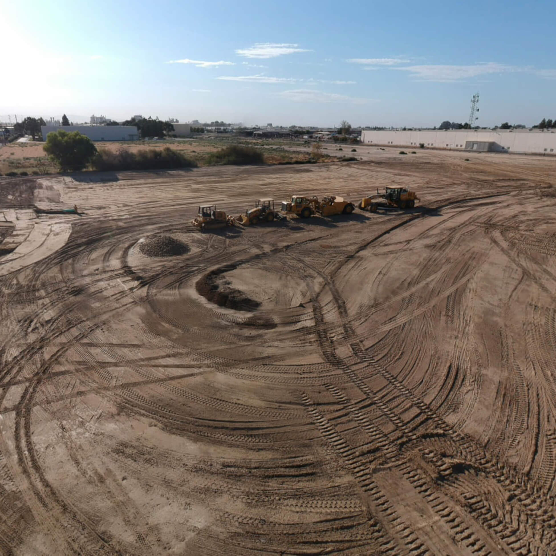 Large cleared construction site with four pieces of heavy machinery and several dirt piles under a blue sky.