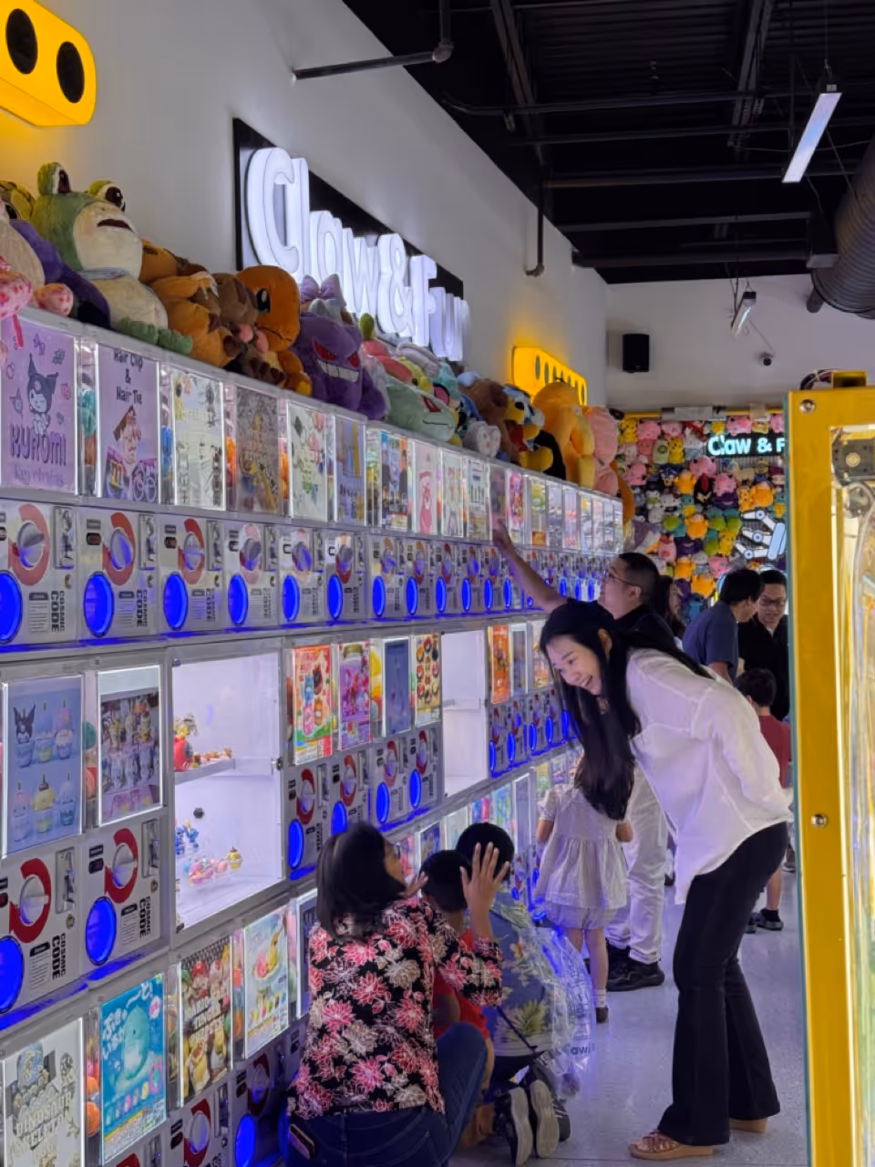People, including children and adults, interact with a row of brightly lit vending machines and colorful plush toys displayed above them in an arcade-style setting.
