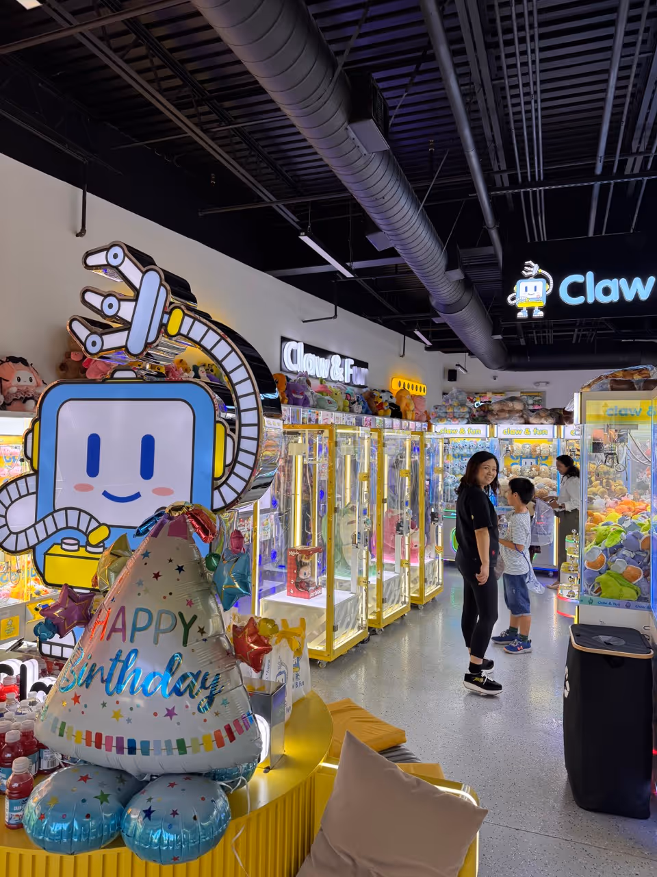 Woman and two children standing inside a brightly lit arcade filled with claw machines and colorful plush toys.