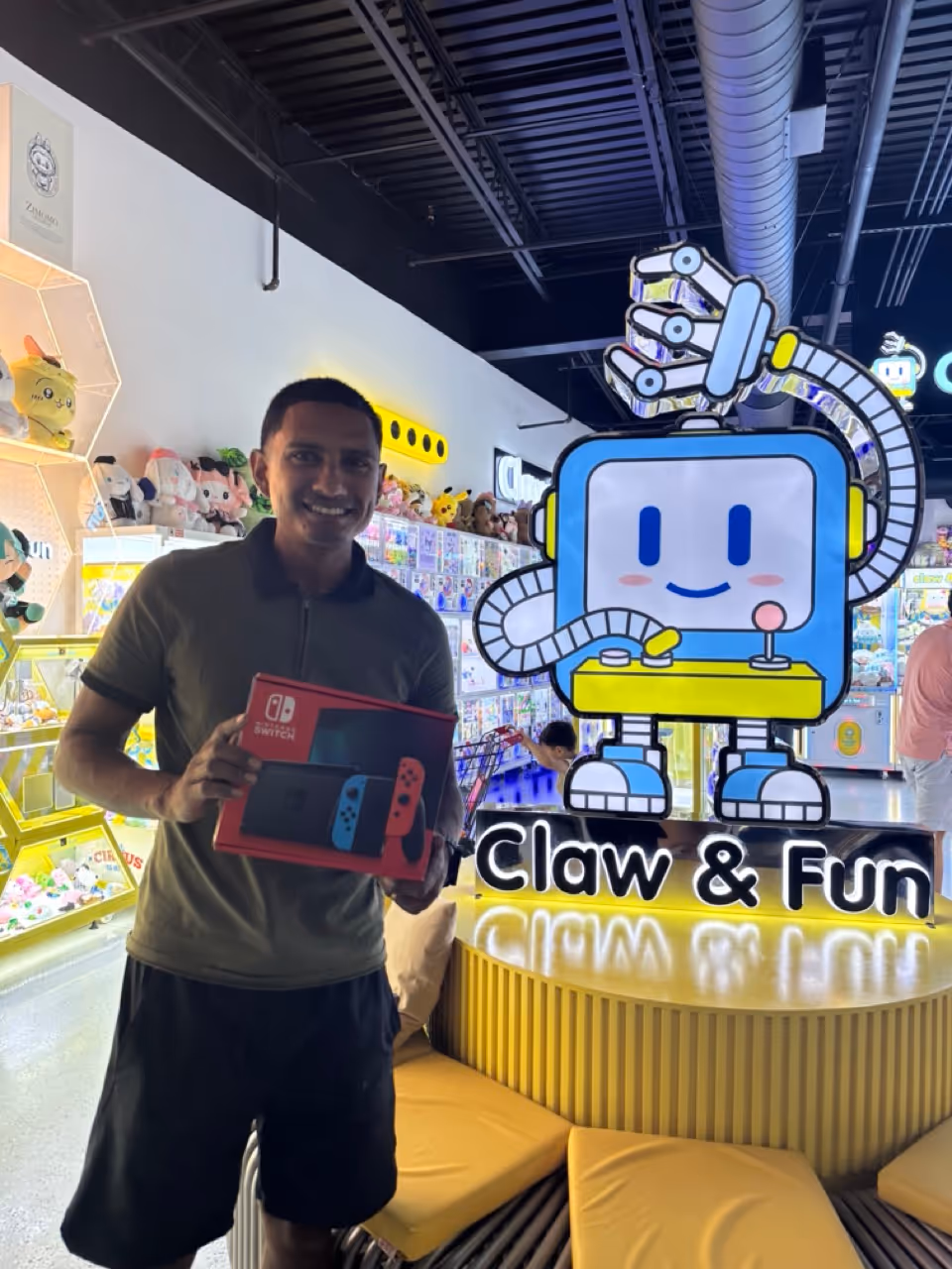 Man smiling and holding a Nintendo Switch box inside a claw machine arcade named Claw & Fun.