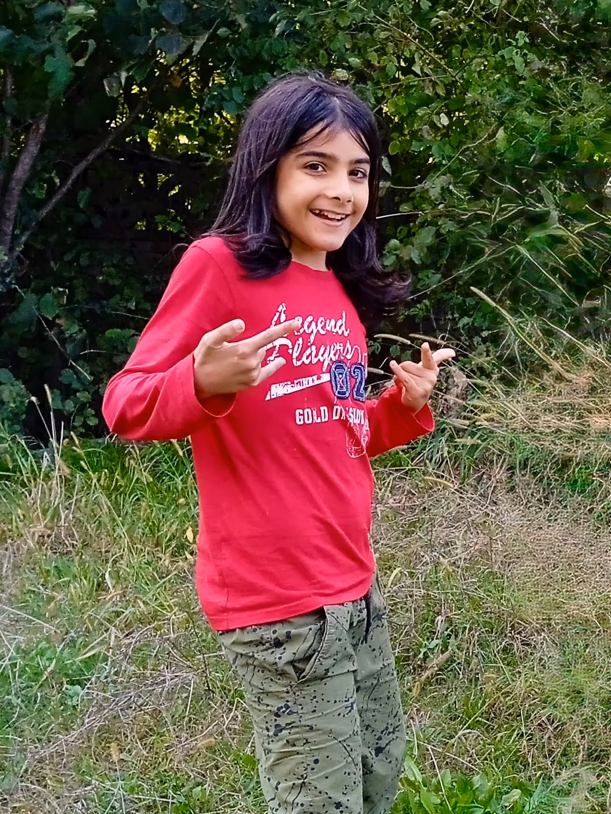 Smiling child in a red long-sleeve shirt and green pants posing outdoors with greenery in the background.