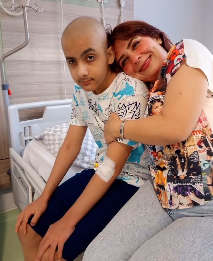 A woman affectionately hugging a young patient with a bald head and an IV line in his arm, seated on a hospital bed.
