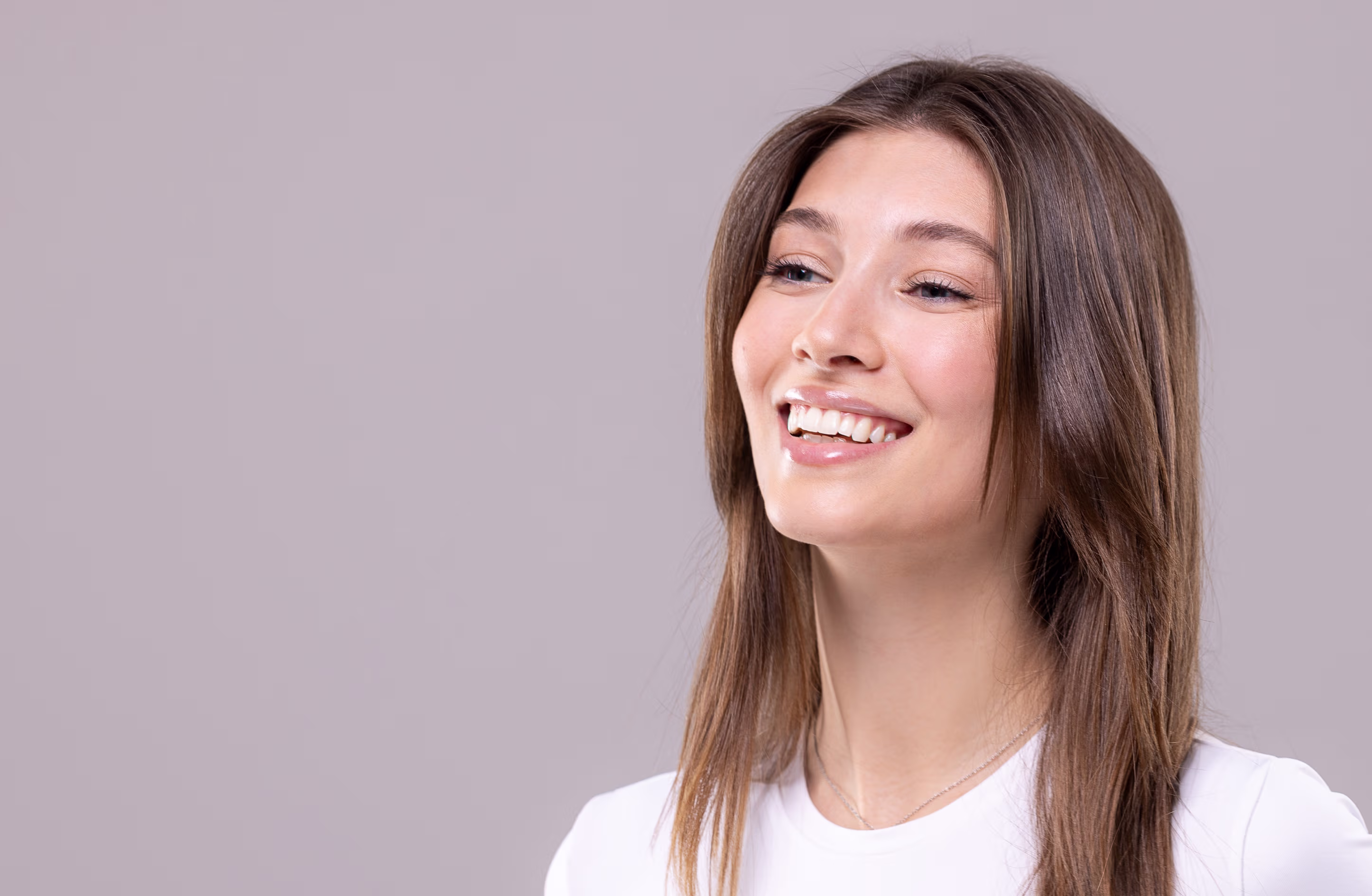 Lächende Frau mit langen braunen Haaren und weißem T-Shirt vor grauem Hintergrund.