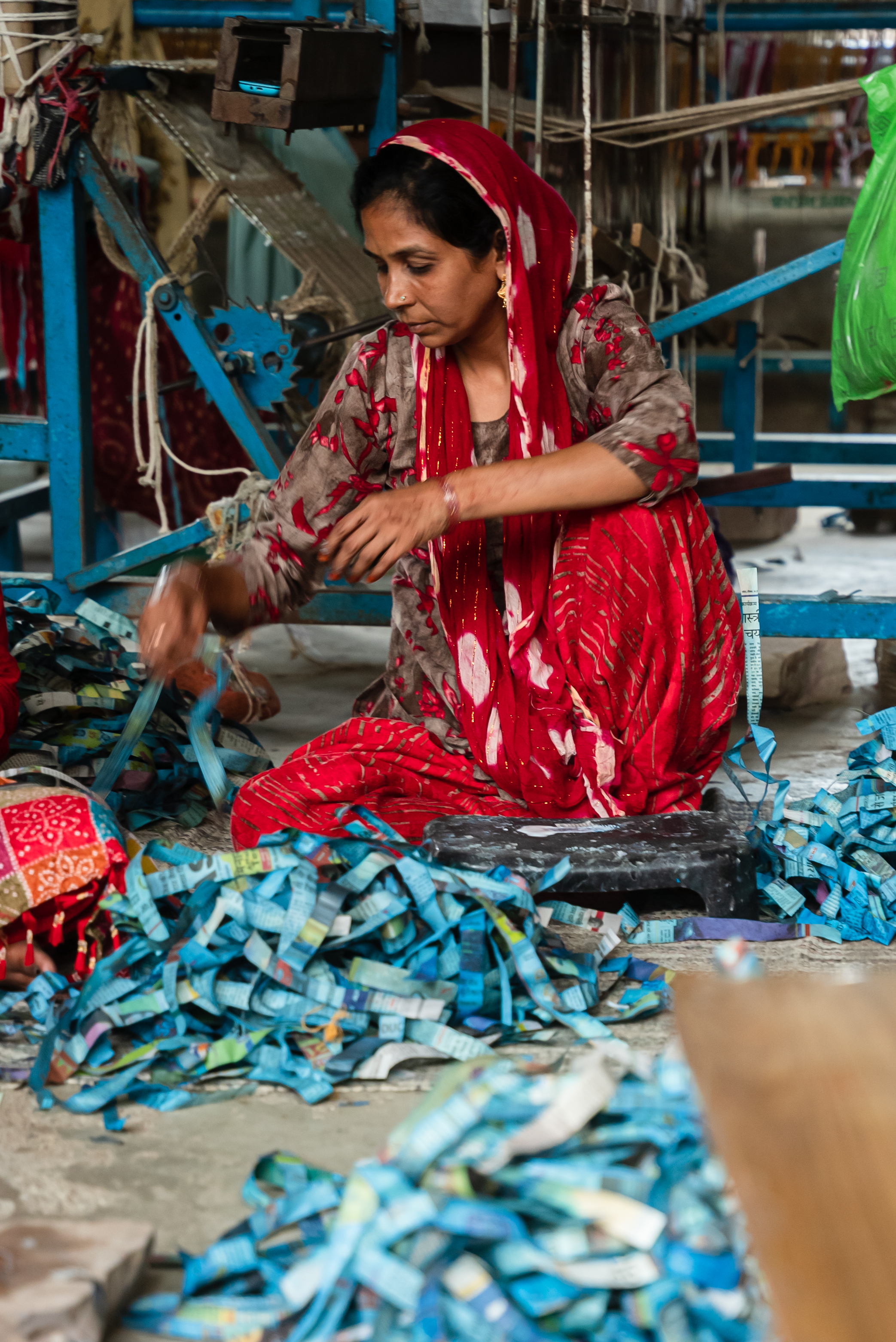 Woman wearing a red and gray patterned outfit sitting on the floor surrounded by blue and multicolored strips of paper, working with her hands.