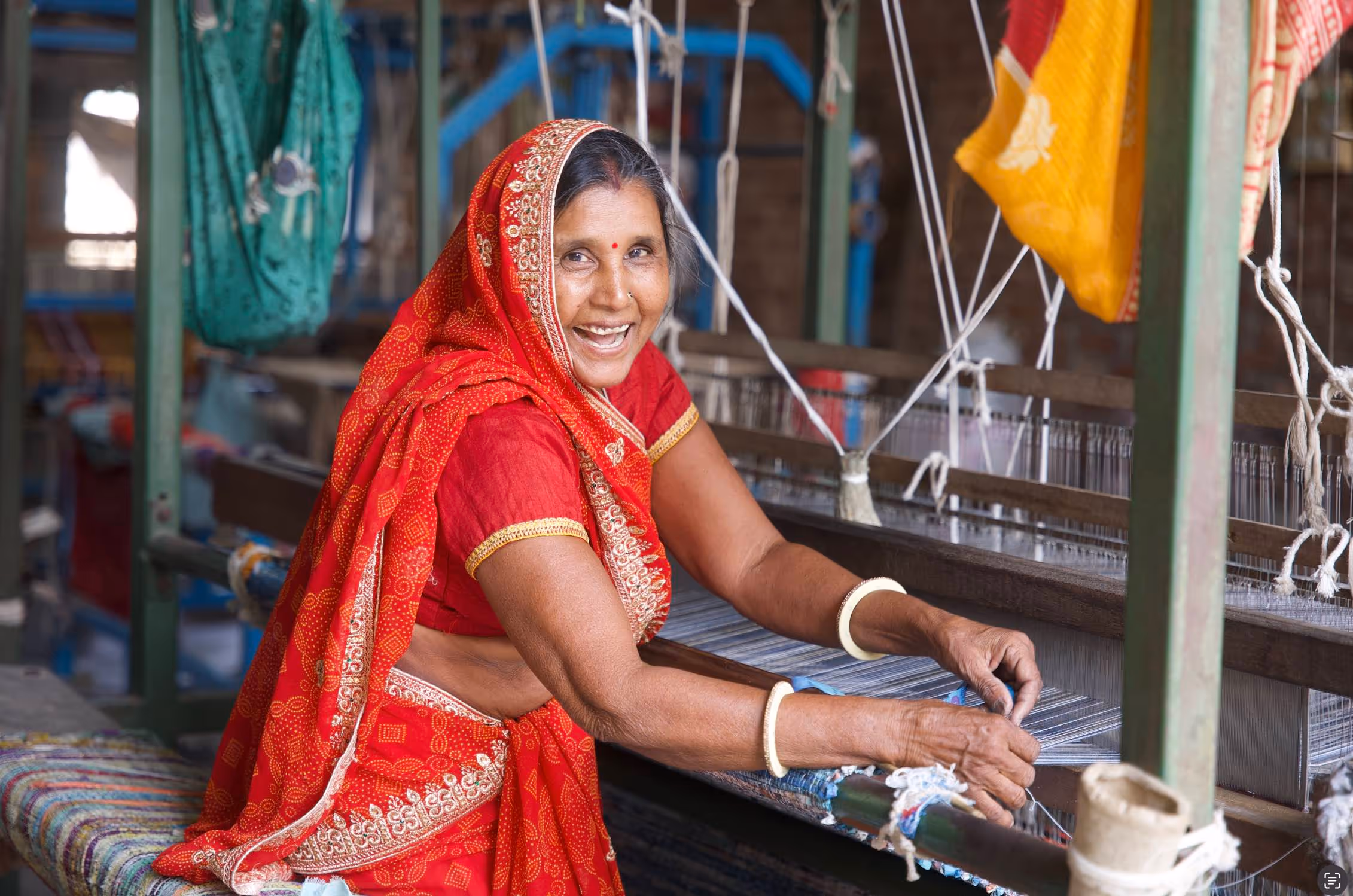 Smiling woman in a red sari weaving fabric on a traditional loom.