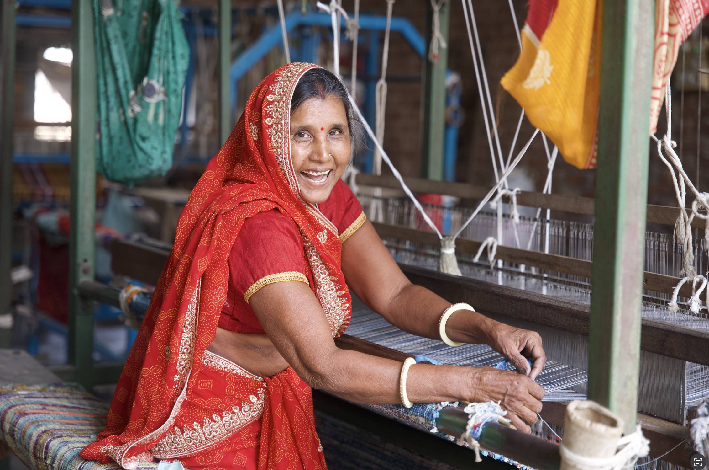 Smiling woman in a red sari weaving fabric on a traditional loom.